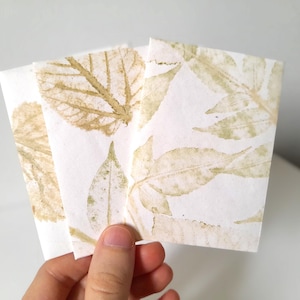 Op de afbeelding: Drie witte kaarten met bladafdrukken in groen- en bruintinten worden vastgehouden. De bladafdrukken variëren in grootte en detail, waardoor een natuurlijk, botanisch thema ontstaat. De kaarten zijn lichtjes op elkaar gestapeld.