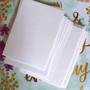 May include: A stack of blank, white rectangular cards. The cards are stacked on a floral patterned background with pink, purple, and gold accents. The cards have a slightly textured surface.
