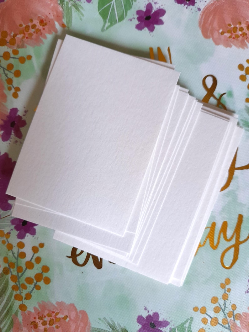 May include: A stack of blank, white rectangular cards. The cards are stacked on a floral patterned background with pink, purple, and gold accents. The cards have a slightly textured surface.