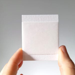 May include: A square, white, absorbent pad with a textured border held between two fingers. The pad is clean and appears to be made of a soft, non-woven material. The background is a solid, light gray.
