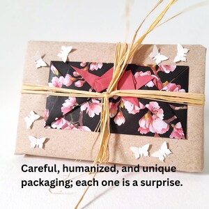 May include: A wrapped gift with a kraft paper exterior, adorned with a floral patterned paper and a red origami crane. The package is tied with raffia and decorated with small white paper butterflies. The text on the image says: Careful, humanized, and unique packaging; each one is a surprise.