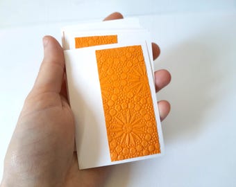May include: A stack of white cards with an embossed orange floral design. The cards are held in a hand, showcasing the textured surface and the bright color of the design. The floral pattern includes circles and starburst shapes.