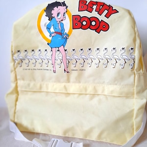 May include: A pale yellow backpack featuring a graphic of Betty Boop in a blue dress inside a yellow circle, with the text BETTY BOOP in red. A horizontal row of white flamingo illustrations runs across the centre, and a white strap with a plastic buckle.