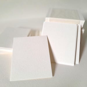 Può includere: Pile di biglietti bianchi quadrati e strutturati. I biglietti hanno una superficie leggermente ruvida, ideale per scrivere o dipingere. L'immagine mostra più pile, con alcuni biglietti a ventaglio. Lo sfondo è una superficie bianca.