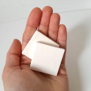Può includere: Due piccoli dischetti cosmetici quadrati bianco sporco sono tenuti in una mano aperta. I dischetti sono impilati, uno sovrapposto leggermente all'altro. Lo sfondo è una superficie bianca uniforme.