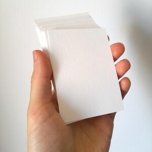 May include: A stack of blank, rectangular, white cards held in a hand. The cards have a slightly textured surface and are stacked neatly. The background is a plain white wall.