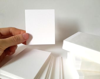 May include: A hand holding a blank white square card, with stacks of similar cards in the background. The cards are a bright white color and have a slightly textured surface. The image is well-lit and the cards appear to be made of paper.
