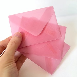 May include: A stack of translucent pink envelopes. The envelopes are square with a flap closure. The envelopes are stacked at a slight angle, with the top envelope partially obscuring the ones below.