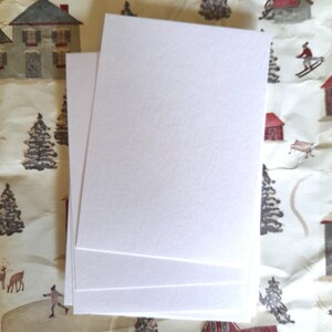 May include: A stack of blank, white rectangular cards. The cards are stacked on top of each other, with a textured surface. The background features a winter-themed pattern with houses, trees, and skiers.