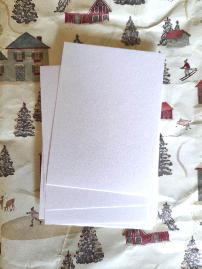 May include: A stack of blank, white rectangular cards. The cards are stacked on top of each other, with a textured surface. The background features a winter-themed pattern with houses, trees, and skiers.