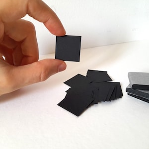 May include: A hand holding a small, black square. Several other black squares are scattered below, with a stack of grey squares on the right. The items are on a white surface.