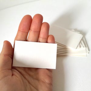 Op de afbeelding: Een hand die een enkele blanco, rechthoekige witte visitekaart vasthoudt, met een stapel vergelijkbare kaarten op de achtergrond. De kaarten zijn effen, met een glad oppervlak, ideaal voor personalisatie of beschrijven.