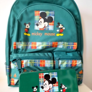 Op de afbeelding: Een donkergroene rugzak en een bijpassend rechthoekig etui met een ruitvormige patch met een Mickey Mouse-afbeelding, twee kleinere Mickey Mouse-figuren en de tekst "mickey mouse" in het oranje gedrukt op beide items.