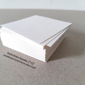 May include: A stack of white square paper sheets, each measuring 2 inches by 2 inches. The edges of the paper are visible, showing the thickness of the stack. Text on the image reads: twinchies blanks 2"x2" onedreamonemirror.etsy.com.