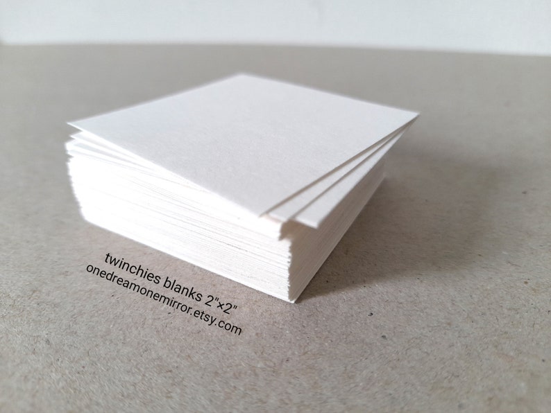May include: A stack of white square paper sheets, each measuring 2 inches by 2 inches. The edges of the paper are visible, showing the thickness of the stack. Text on the image reads: twinchies blanks 2"x2" onedreamonemirror.etsy.com.