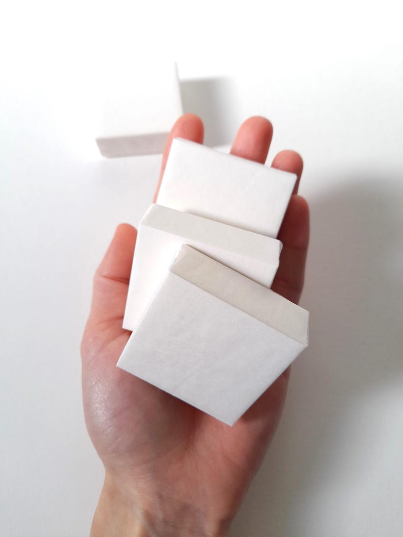 May include: A hand holding several small, white, square-shaped paper boxes. The boxes are stacked and slightly angled, with a strip of tape visible on the top edge of one. The background is plain white.