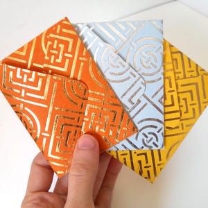 May include: A hand holds a stack of colorful, square envelopes. The envelopes are orange, light blue, and yellow, each with a gold geometric pattern. The envelopes are made of paper and are likely for cards or small gifts.