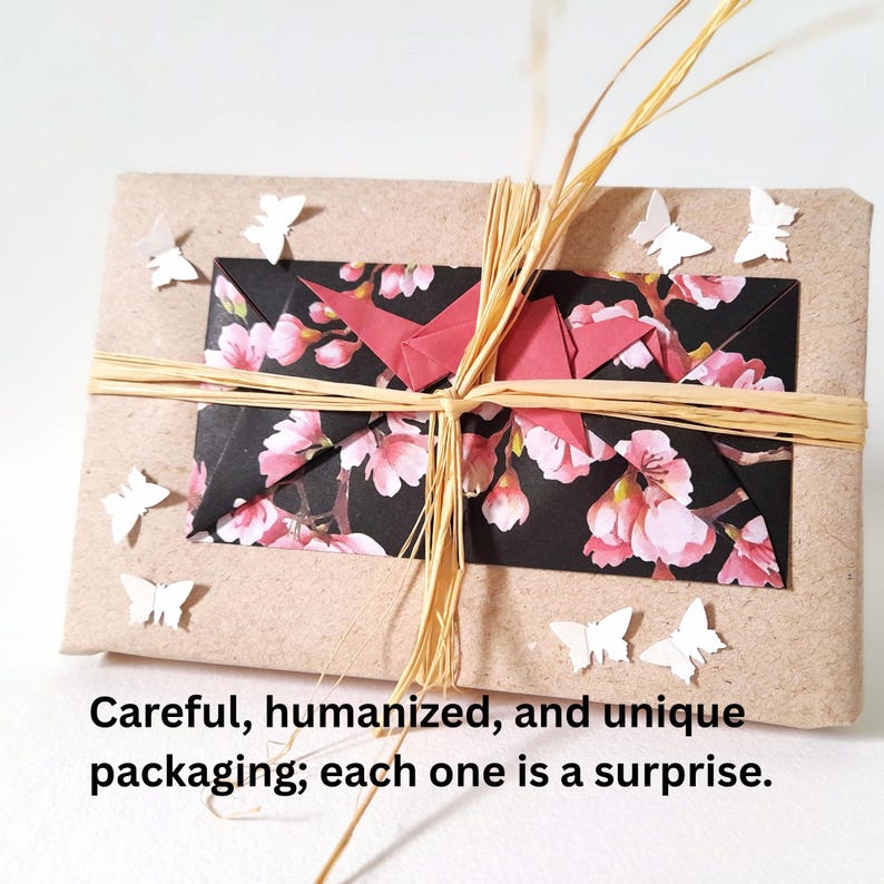 May include: A wrapped gift with a kraft paper exterior, adorned with a floral patterned paper and a red origami crane. The package is tied with raffia and decorated with small white paper butterflies. The text on the image says: Careful, humanized, and unique packaging; each one is a surprise.