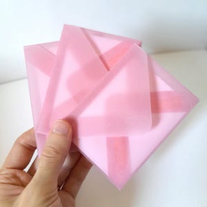May include: A stack of square, translucent pink vellum envelopes. The envelopes are layered, showing a soft, semi-transparent quality with a smooth, matte texture. Each envelope features a triangular flap folded over the center of the square base.
