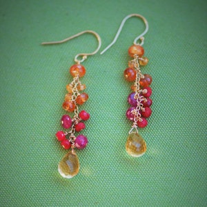 May include: A pair of gold earrings with a teardrop-shaped gemstone at the bottom. The earrings are made with a chain of small orange, red, and pink gemstones.