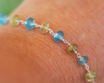 May include: A silver bracelet with blue and green gemstone beads. The bracelet is on a person's wrist.