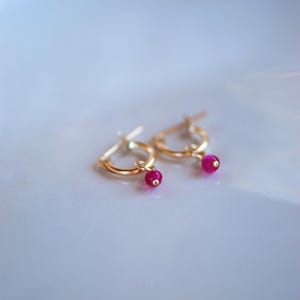 May include: A pair of gold hoop earrings with a small, dangling pink gemstone on each earring.