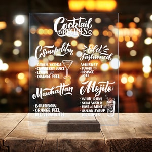 May include: A clear acrylic sign with white text listing cocktail recipes. The sign has the title "Cocktail Recipes" and lists the ingredients for a Cosmopolitan, Old Fashioned, Manhattan, and Mojito.
