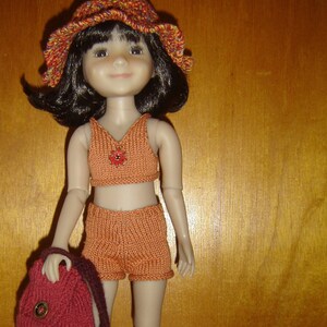 Peut inclure: Une poupée portant une tenue tricotée orange et marron avec un chapeau assorti. La poupée tient un sac rouge tricoté. La poupée est debout sur une surface en bois avec un rocher brun au premier plan.