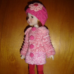 Peut inclure: Un manteau en tricot rose pour poupée avec une fleur rose sur le chapeau. La poupée porte un pantalon rouge et des bottes noires.