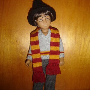 Peut inclure: Une poupée portant un chapeau pointu noir, un pull gris, un pantalon gris et une écharpe rayée rouge et or.