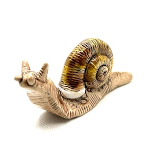 Könnte beinhalten: Keramik-Schneckenfigur mit detailliert strukturierter Schale in Braun- und Gelbtönen. Der Körper der Schnecke ist hellbeige, mit einem weißen Band um die Basis der Schale. Die Figur hat zwei kleine Fühler und ist vor weißem Hintergrund platziert.
