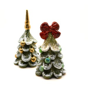 Pode incluir: Duas árvores de Natal de cerâmica, uma com um laço vermelho e a outra com um topo dourado. Ambas as árvores são decoradas com enfeites verdes e dourados e têm um acabamento branco nevado.
