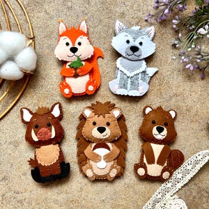 May include: A collection of handmade felt woodland animal ornaments, including a fox, wolf, boar, hedgehog, and bear. Each ornament is intricately detailed with various colors and textures, perfect for seasonal decor or children's crafts.