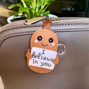 Może przedstawiać: Brązowy brelok z filcu z uśmiechniętą buzią i napisem "Wierzę w ciebie" na białym znaku. Brelok jest przymocowany do brązowej skórzanej torebki.