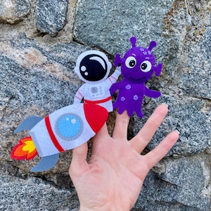 Op de afbeelding: Een set van drie vingerpoppen van vilt met een astronaut in een raket, een paarse alien en een hand die alle drie poppen draagt.