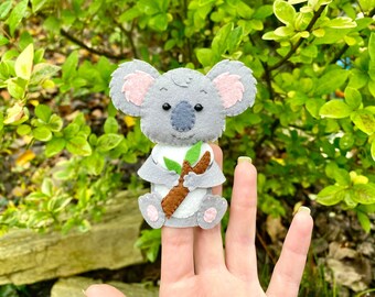 Può includere: Un pupazzo per dita di koala in feltro grigio che tiene un bastone marrone con foglie verdi. Il pupazzo ha orecchie rosa e occhi neri.