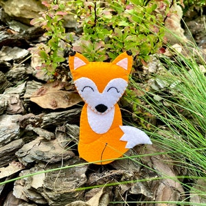 May include: A felt fox toy with orange fur, white chest, and a black nose. The toy is sitting on a bed of brown wood chips and green grass.