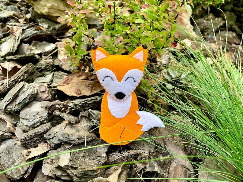 May include: A felt fox toy with orange fur, white chest, and a black nose. The toy is sitting on a bed of brown wood chips and green grass.
