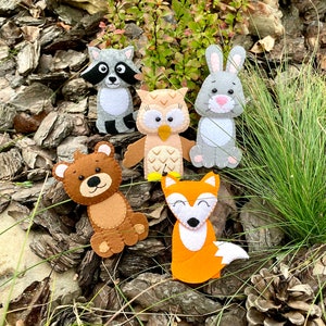 May include: Five felt finger puppets depicting woodland animals: a raccoon, an owl, a rabbit, a bear, and a fox. The puppets are brown, gray, orange, and white.