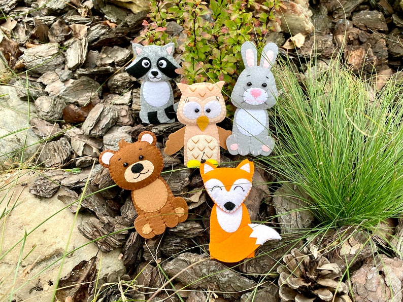 May include: Five felt finger puppets depicting woodland animals: a raccoon, an owl, a rabbit, a bear, and a fox. The puppets are brown, gray, orange, and white.
