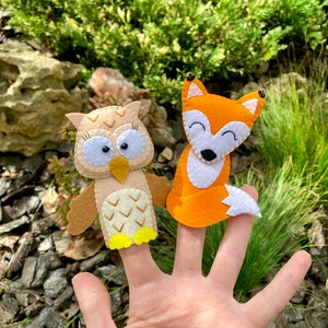 May include: Two felt finger puppets, an owl and a fox, are worn on a person's fingers. The owl is brown with white accents and yellow feet. The fox is orange with white accents.