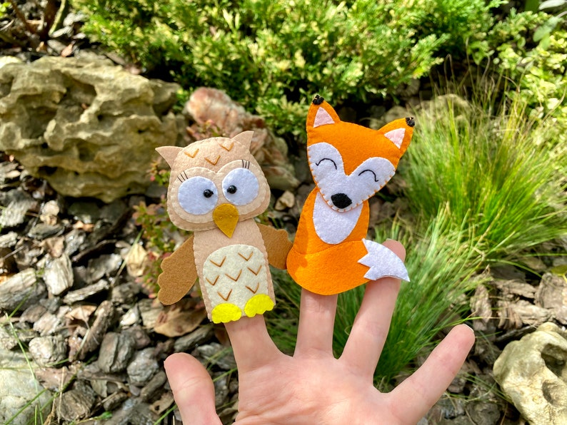 May include: Two felt finger puppets, an owl and a fox, are worn on a person's fingers. The owl is brown with white accents and yellow feet. The fox is orange with white accents.