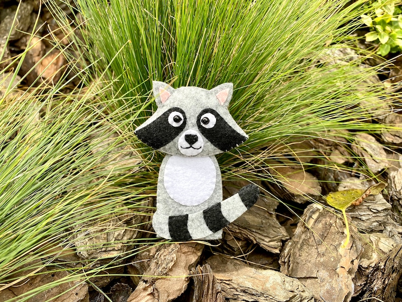 May include: A gray and black felt raccoon finger puppet with white eyes and a white belly. The puppet is sitting on a bed of brown wood chips and green grass.
