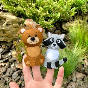 May include: Two felt finger puppets, a brown bear and a gray raccoon, are worn on a person's fingers. The puppets are made of felt and have embroidered details.