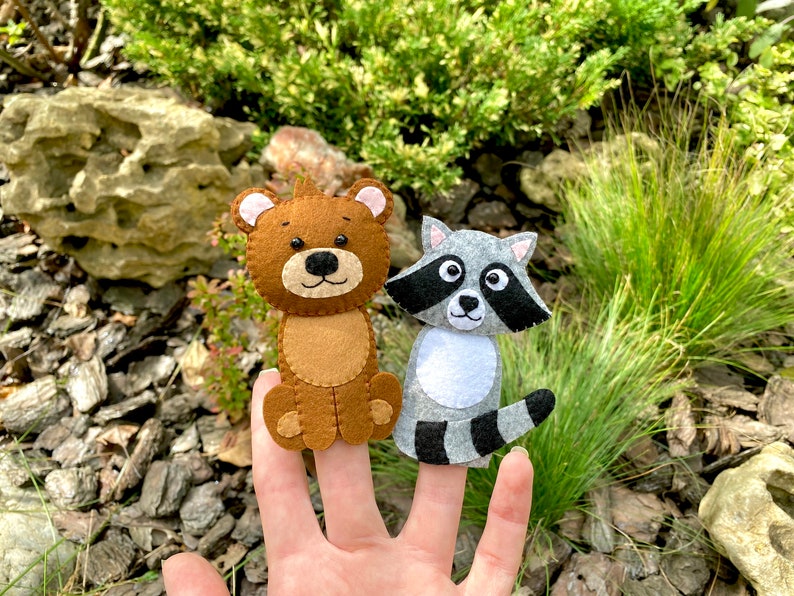 May include: Two felt finger puppets, a brown bear and a gray raccoon, are worn on a person's fingers. The puppets are made of felt and have embroidered details.