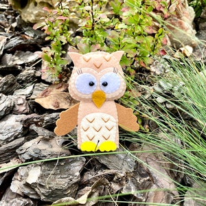 May include: A brown felt owl with large black eyes and a yellow beak. The owl is sitting on a bed of brown wood chips.