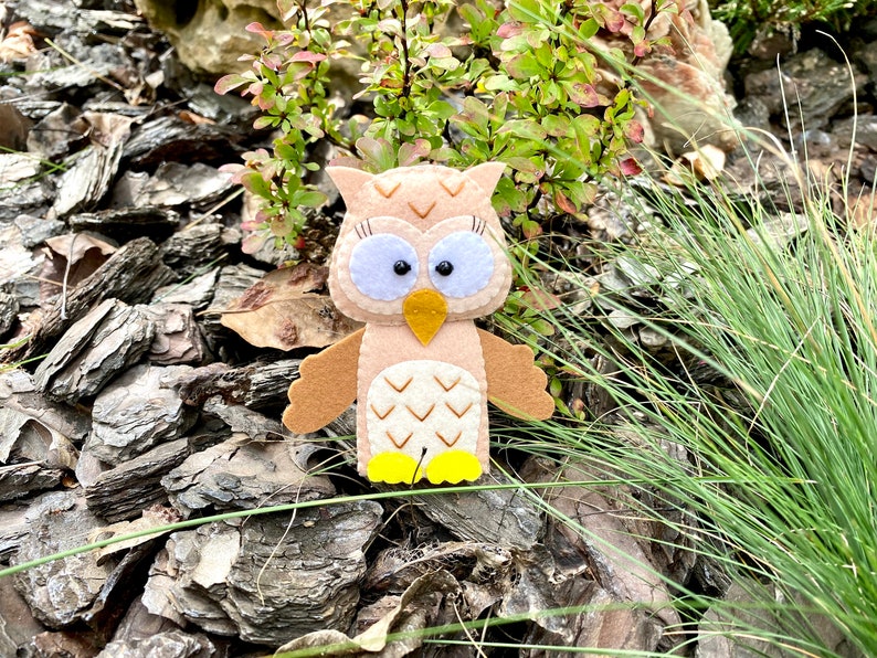May include: A brown felt owl with large black eyes and a yellow beak. The owl is sitting on a bed of brown wood chips.