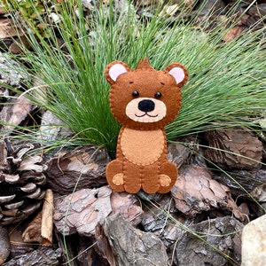 May include: A brown felt bear with pink ears and black eyes. The bear is sitting on a bed of brown pine bark.