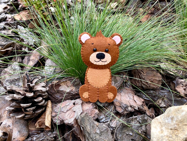 May include: A brown felt bear with pink ears and black eyes. The bear is sitting on a bed of brown pine bark.
