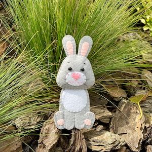 May include: A gray felt bunny with pink ears and nose, sitting on a bed of brown wood chips.